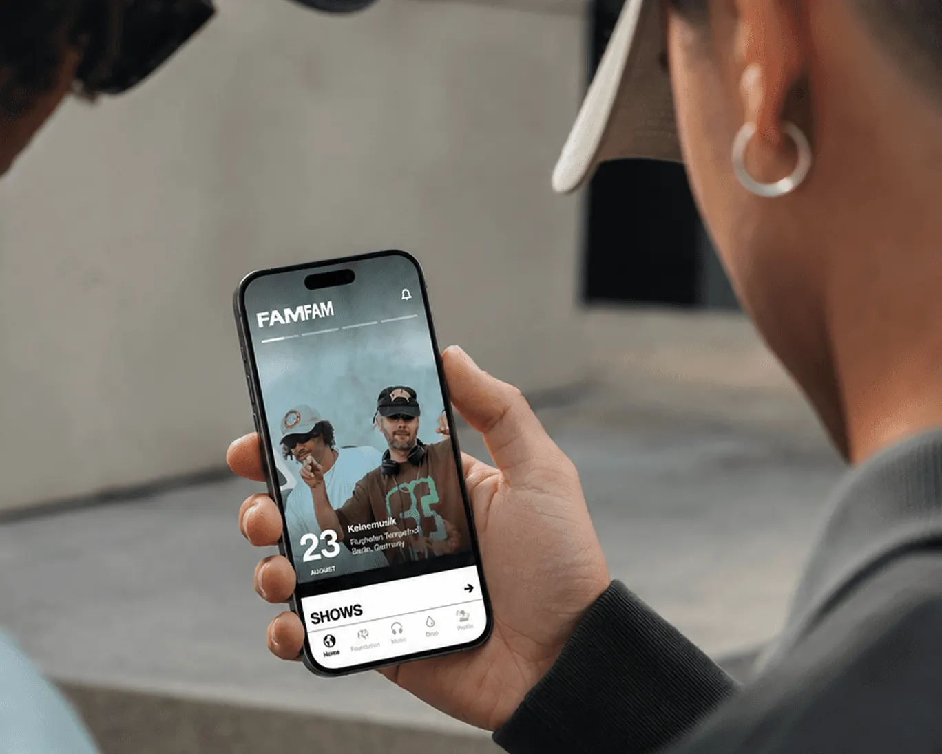 Hand haelt ein Smartphone mit der FAMFAM-App, im Hintergrund zwei Personen
