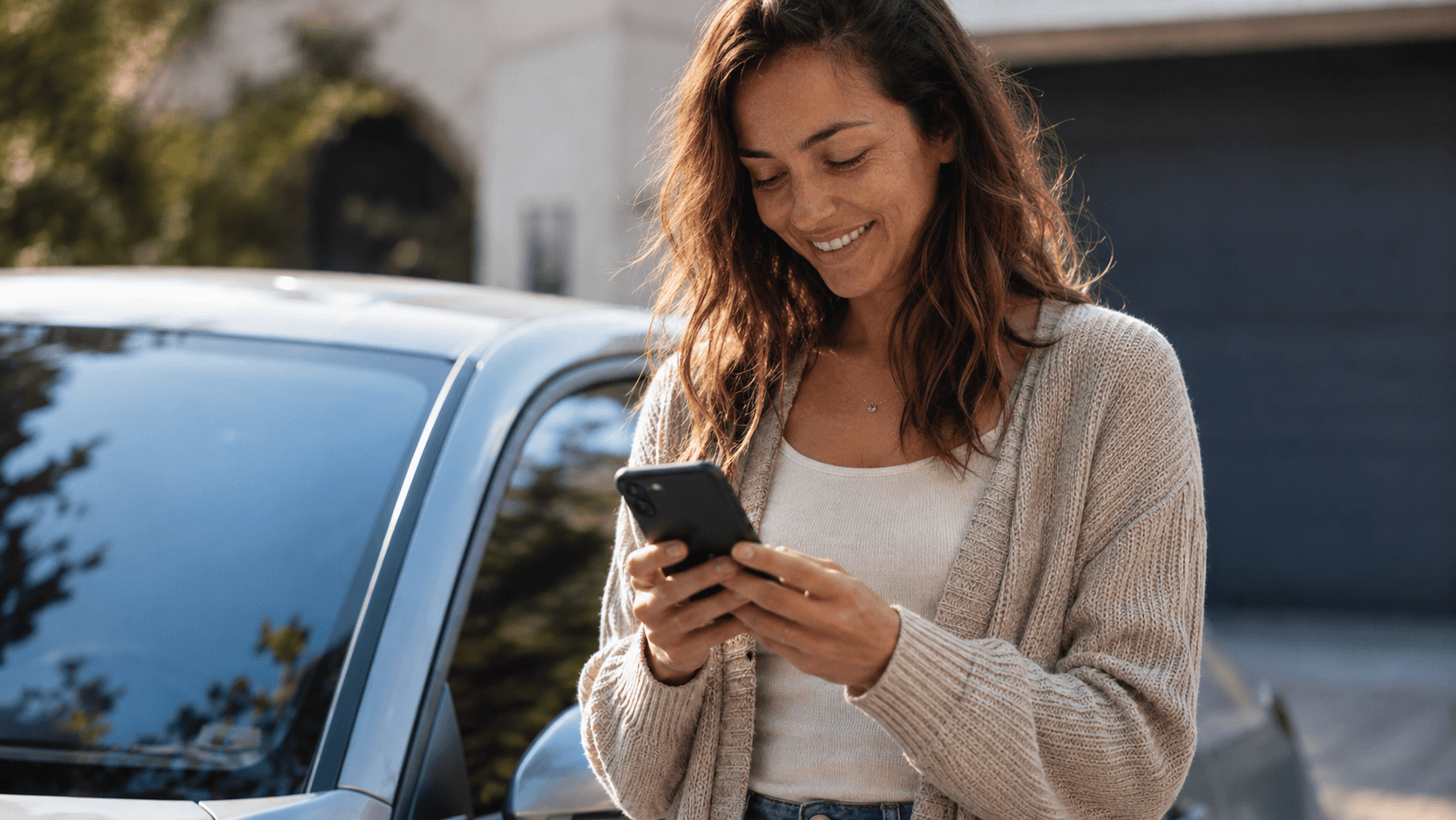 Frau lehnt lächelnd an einem hellblauen Auto und schaut auf ihr Smartphone.