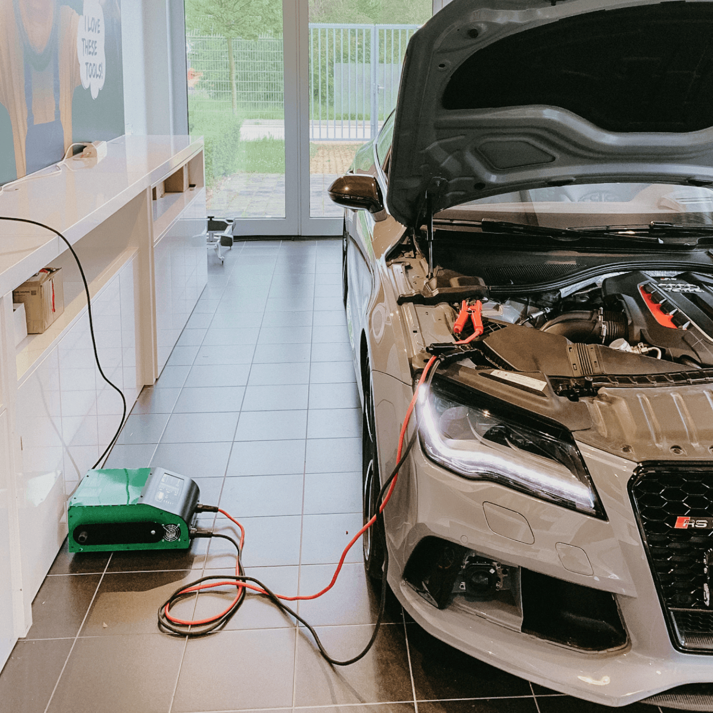 Workshop with a car, bonnet open, its battery connected by red and black cables to a support unit on the floor