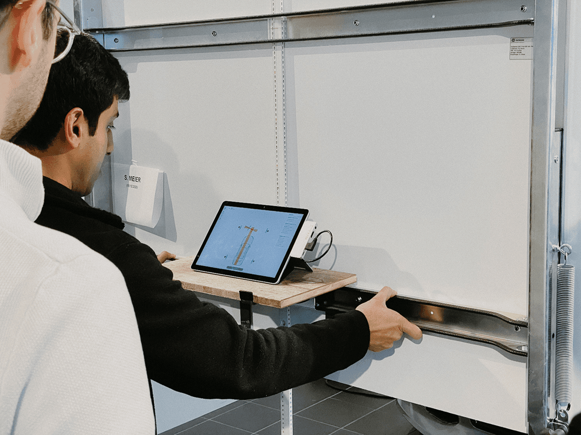 A man in front of a calibration panel looks at a tablet showing a technical drawing and points at the mounting rail below