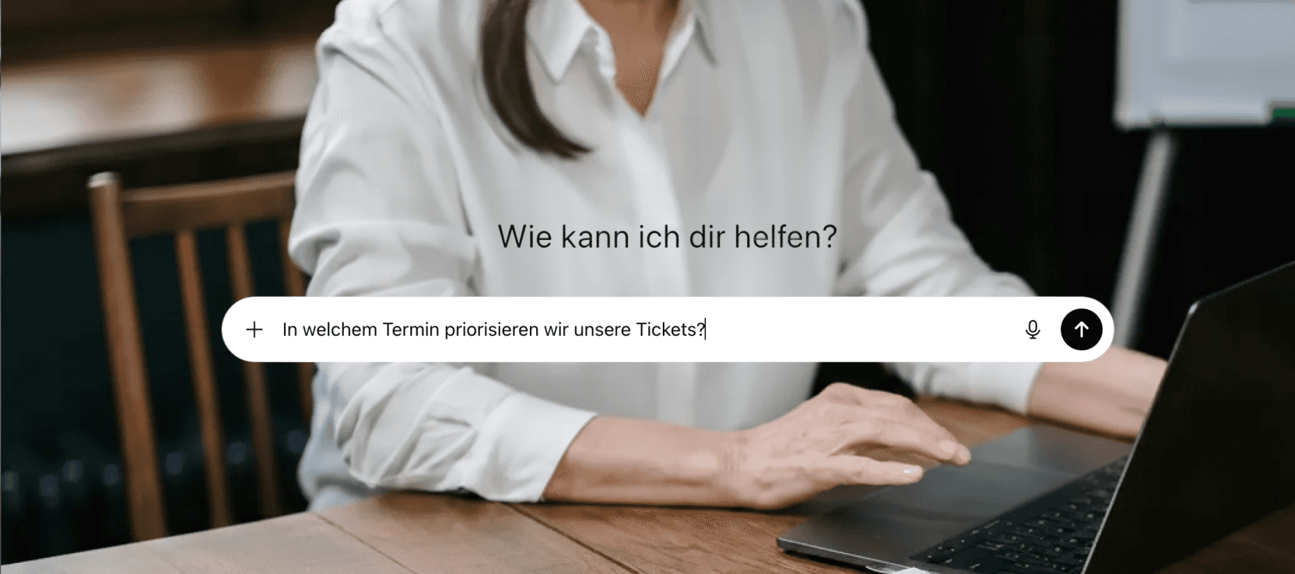 Frau am Laptop, davor ein Eingabefeld mit der Frage „Wie kann ich dir helfen?“ und einer eingetippten Frage zur Ticketpriorisierung