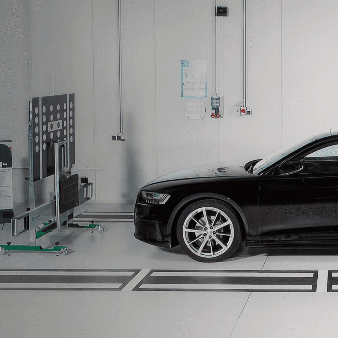 Bright workshop: a black car stands beside a mobile calibration panel, with alignment markings set into the floor