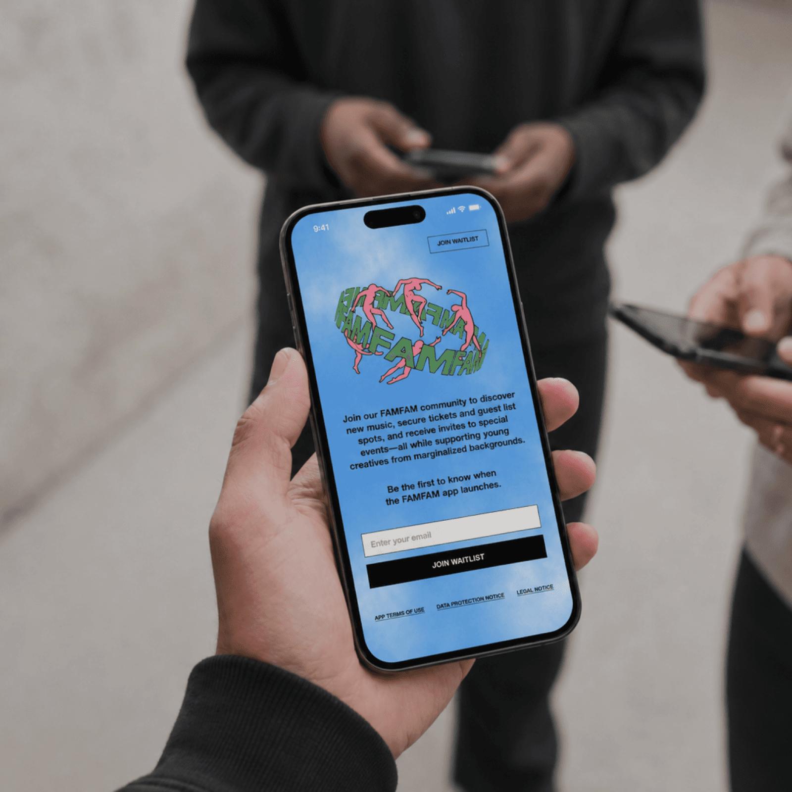 Eine Hand haelt ein Smartphone mit dem FAMFAM-Anmeldebildschirm fuer die Warteliste, im Hintergrund zwei Personen an ihren Telefonen