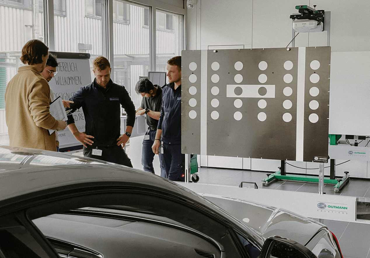 Five people stand around a calibration panel with a dot pattern in a workshop, two vehicles in the foreground